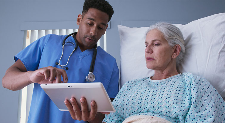 Nursing student showing a patient in a hospital bed information on a tablet